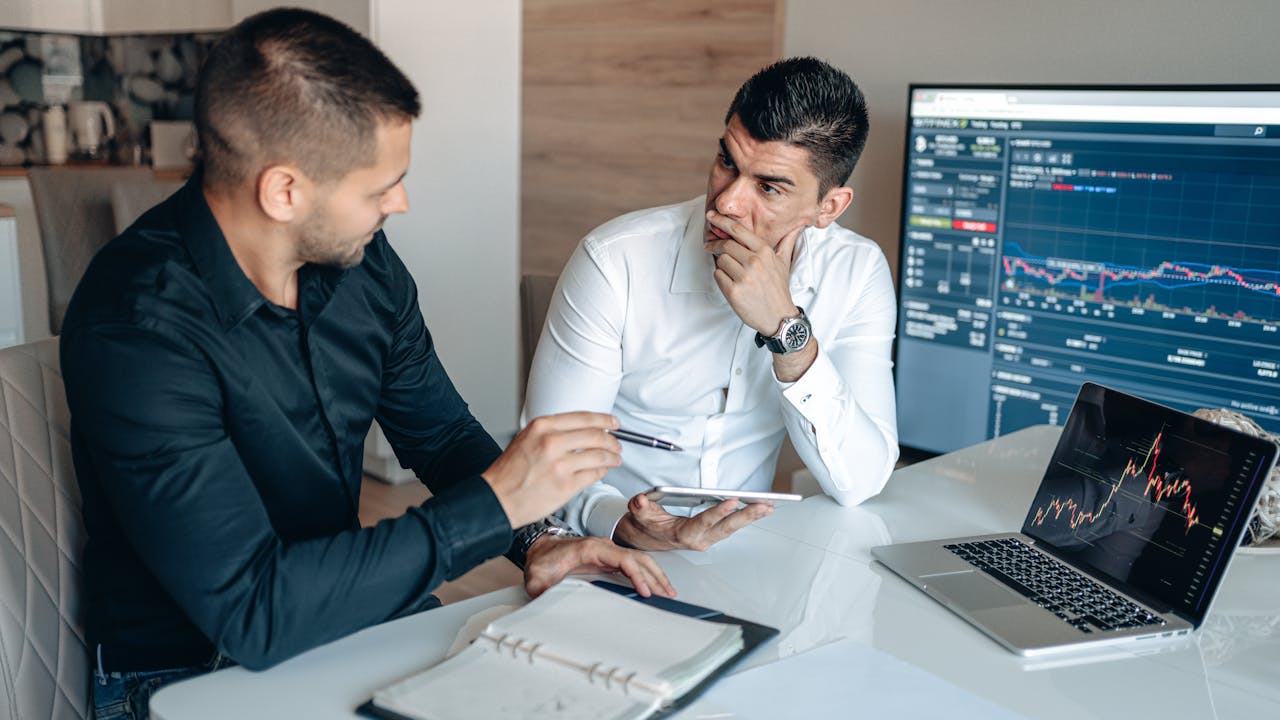 Two businessmen discussing stock market trends with trading screens in an office setting.